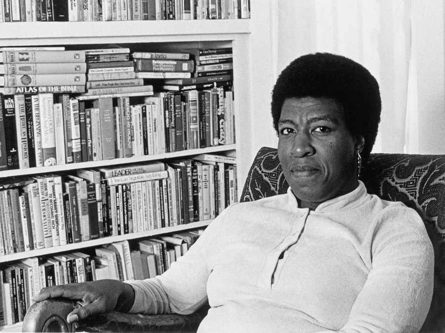 A black and white photograph of Octavia Butler seated at home in front of several bookshelves.