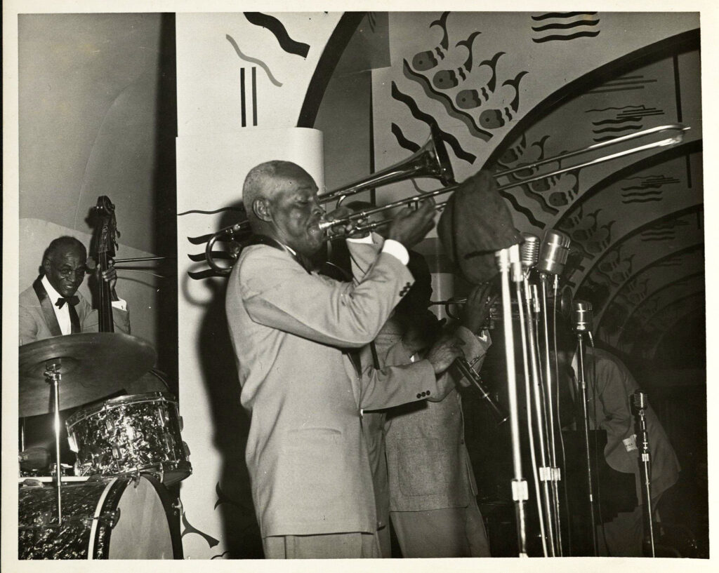 A band featuring l. to r.: Richard McLean, bass; Bill Matthews, trombone; unidentified clarinet player; Alvin Alcorn, trumpet; and Joe Robichaux, piano, perform during Jazz Fest’s Steamboat Stomp evening concert on the Steamer President riverboat, April 18, 1974, photographer: Jerry Bray, Mina Lea Crais collection HJA-060, 117.099, Tulane University Special Collections.