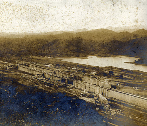Aerial view of a large construction site with a body of water and mountains in the background.