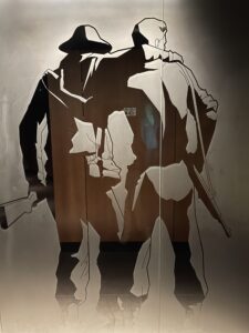 Photo of two soldiers from the back one wearing a hat and one not. Both are holding guns to their side and dressed in army uniforms